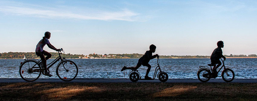 Barn som cyklar på olika typer av cyklar längs med sjö